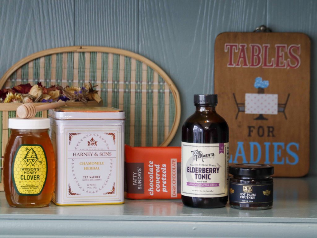 A jar of Wixson's honey, tin of Harney & Sons chamomile herbal tea, Fatty Sundays dark chocolate-covered pretzel rods, elderberry tonic by Food & Ferments, and Beth's Farm kitchen hot plum chutney, all lined up on a shelf.